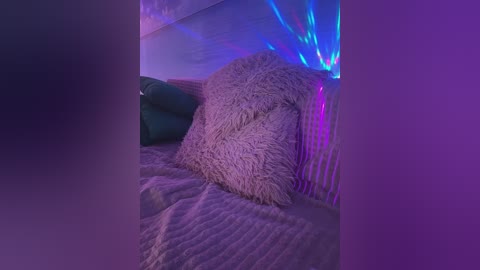 Media: Video of a plush, gray, shaggy pillow on a bed with a textured, light gray blanket, against a purple and blue LED-lit wall. The room exudes a cozy, modern, and slightly futuristic ambiance.