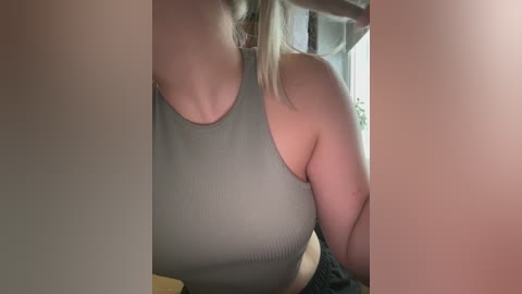 Video of a fair-skinned woman with blonde hair, wearing a gray ribbed tank top, captured from the side, partially obscured by out-of-focus objects.