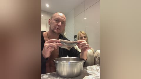 Media: Video of a bald, muscular man holding a fish in a modern kitchen with a blonde woman in the background.