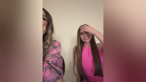 Media: Video of two young women, one with long, wavy blonde hair, wearing a pink plaid shirt, partially hidden by a white wall; the other with straight, long brown hair, smiling, wearing a pink top, holding her hair.