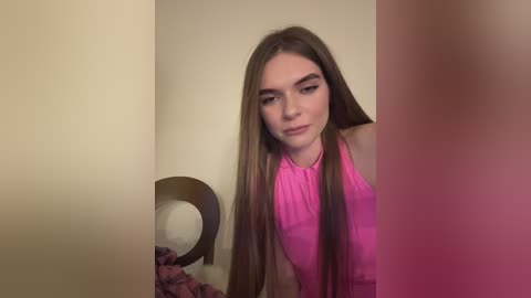 Media: Video of a young Caucasian woman with long brown hair and fair skin, wearing a pink halter top, standing in a dimly lit room with beige walls and a wooden chair.