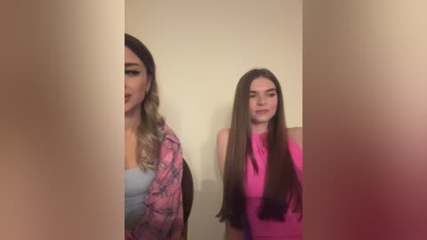 Media: Video of two young women with long, straight hair. One wears a pink top, the other a floral jacket. Both stand against a beige wall, with a blurry foreground.