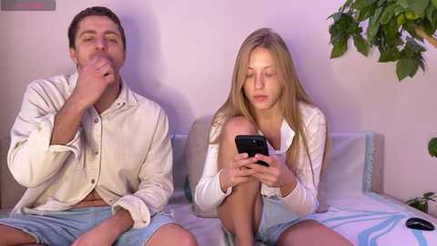 Media: Video of a blonde woman in a white top and blue shorts, looking at a smartphone, next to a man in a white shirt and blue shorts, yawning, in a minimalist bedroom.