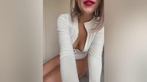 Video of a young woman with light skin and shoulder-length, platinum blonde hair, wearing a white, long-sleeve, plunging neckline top, sitting in a beige room.