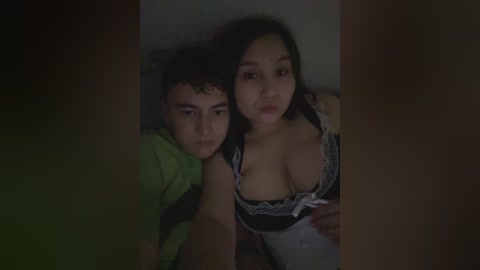 Media: Video of a young couple lying in bed, dimly lit. The man, with curly hair, wears a green shirt; the woman, with dark hair, is in a black and white lingerie top, showcasing cleavage.
