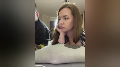 Media: Video of a young woman with long, light brown hair, wearing a black and white patterned top, resting her chin on her hand in a thoughtful pose.