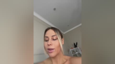Video of a topless woman with light skin and blonde hair, eyes closed, smiling, standing in a modern, minimalist room with beige walls and white shelves holding decorative items.
