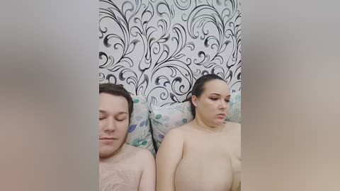 A video of a nude, light-skinned couple lying side by side on a bed. The man has a light beard and the woman has shoulder-length dark hair, both with neutral expressions.