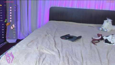 A video of a messy bedroom with a beige bedspread, a plush white dog toy, and a black remote control on the bed. Neon purple lighting on the wall, partially visible on the left.