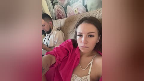 Media: Video of a young woman in a pink shirt and necklace, lying on a beige sofa, texting, while a man in a hoodie sleeps behind her. Background features floral cushions.