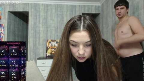 Video of a young woman with long brown hair, wearing a black top, leaning forward, and a shirtless man standing behind her in a room with grey-patterned wallpaper and a purple nightstand with a golden statue.