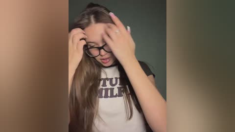 Media: A video of a young woman with long, straight brown hair, wearing glasses, adjusting them, and a white t-shirt with \"TOO MUCH MILK\" printed in black.