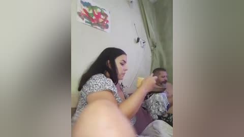 Video of a young woman with long black hair in a patterned top, playing with a young boy in a striped shirt, in a hospital room with green walls and a colorful painting.