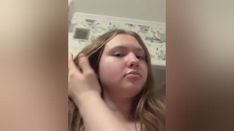 Video of a young Caucasian woman with long, wavy blonde hair, adjusting her hair in a bathroom with floral wallpaper and a white ceiling.
