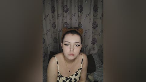 Media: Video of a young Asian woman with light skin, wearing a leopard-print top and brown cat ears, leaning on a bed with floral-patterned curtains in the background.