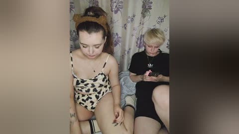 Media: Video of two young women: one in a leopard-print leotard, the other in a black hoodie, sitting on a bed in a room with floral curtains.