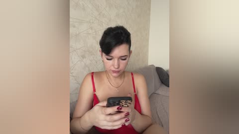 A video of a young woman with short, dark hair, wearing a red spaghetti strap dress, holding a leopard-print phone, seated on a beige couch with a textured wall behind her.