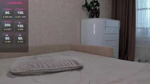 Video of a dimly lit, modern bedroom with a white bed, white dresser, and a potted plant on a windowsill. A digital clock on the wall displays the time and weather.