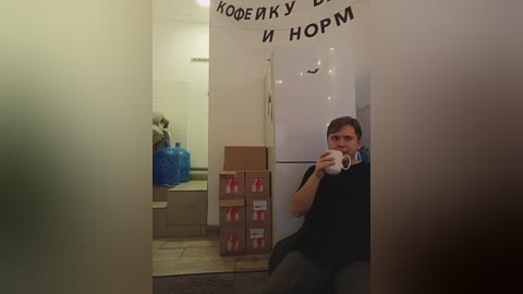 Media: A video captures a man with short brown hair, wearing glasses and a black shirt, taking a selfie in a dimly-lit room. The background shows stacked boxes and blue water containers, with Russian Cyrillic text on a whiteboard.