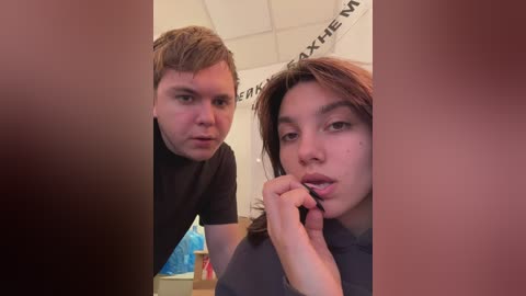 Media: Video of a young woman with long brown hair and a man with short brown hair, both wearing black t-shirts, standing in a dimly lit room with a blue banner in the background.