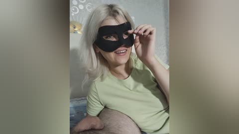 Video of a blonde woman in a black mask, sitting on a bed with a light green shirt, and a man's hairy legs.
