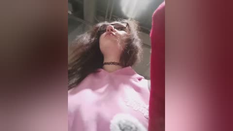 Video of a woman with curly hair, wearing glasses and a pink coat, lying on her back, eyes closed, surrounded by blurred red and pink objects.