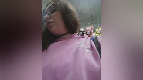 Video of a woman with glasses, brown hair, and a black choker, wearing a pink robe, standing in a dimly lit room with shelves filled with items in the background.