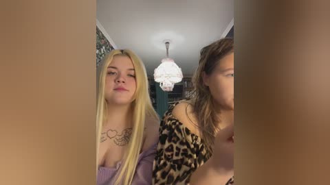 Video of two women in a dimly lit room; one with long blonde hair, purple top, and heart tattoo; the other with brown hair, leopard print top.