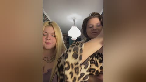 Video of two young women, one blonde with a heart tattoo, and one brunette, wearing leopard print, hugging in a dimly lit room with a chandelier.