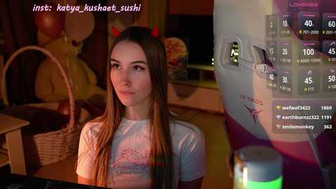 Media: Video of a young woman with long brown hair, wearing a light gray t-shirt and red devil horns, sitting in a cozy, dimly lit room with a computer displaying game statistics in the background.