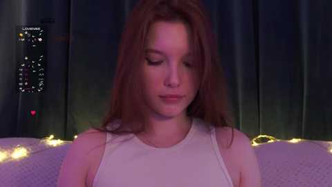 Video of a young woman with fair skin and long red hair, wearing a white tank top, sitting against a dark curtain backdrop with soft yellow lights.