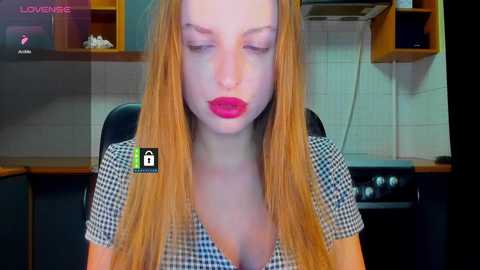 Video of a fair-skinned, blonde woman with full lips, wearing a low-cut checkered top, sitting in a kitchen with tiled walls and wooden cabinets.