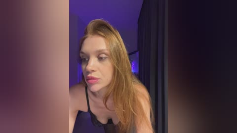 Video of a young, fair-skinned woman with long, straight red hair, wearing a black lace bra, looking pensively at a mirror in a dimly lit room with purple and blue lighting.