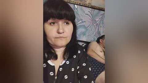 Video of a middle-aged woman with straight black hair and a serious expression, wearing a black dress with white lipstick prints. Behind her, a man with a shaved head is shirtless in blue patterned shorts, sitting on a bed with floral wallpaper.