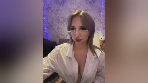 Media: Video of a young woman with light skin, platinum blonde hair, and large glasses, wearing a white button-up shirt, reclining on a chair in a dimly lit room with purple LED strip lighting.