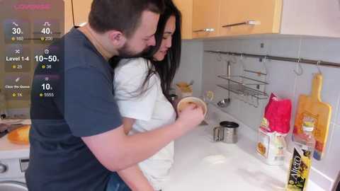 A video depicts a bearded man in a dark T-shirt and a woman in a white shirt standing closely in a modern kitchen with beige cabinets and a white countertop.