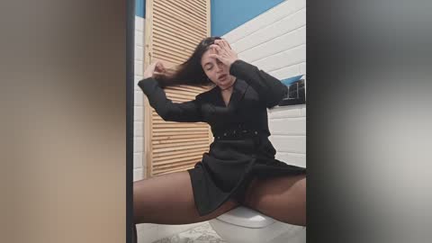 Media: Video of a woman in a black dress, sitting on a toilet, legs spread, hand on head, in a tiled bathroom with wooden blinds.