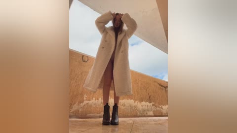 Media: Video of a woman standing on a tiled floor, wearing a long, cream coat, black boots, and a dark shirt, with hands raised, under a white archway, beige wall, and cloudy sky background.