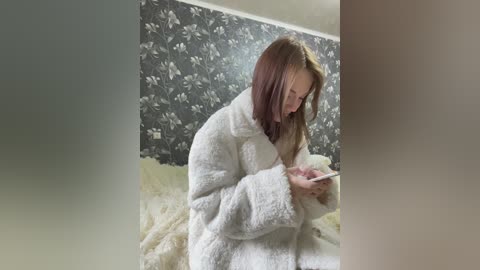 Media: Video of a young woman with straight, shoulder-length auburn hair, wearing a white, plush robe, seated on a beige bed. She's focused on a pink smartphone. The background features a dark floral wallpaper.