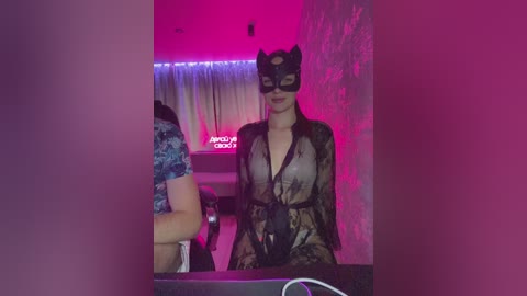 Video of a fair-skinned woman with long black hair, dressed in a sheer black lace gown, wearing a black cat mask, standing in a dimly lit room with pink and purple lighting, and a partially visible person in the background.