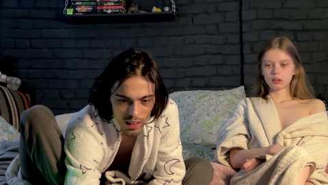 Media: Video of a dark-haired man in a white robe on a bed, a blonde woman in a beige robe beside him, against a brick wall background.