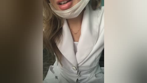 Media: Video of a woman with long, wavy brown hair partially covered by a white surgical mask, wearing a white lab coat and a white top, with a blurred brick wall background.