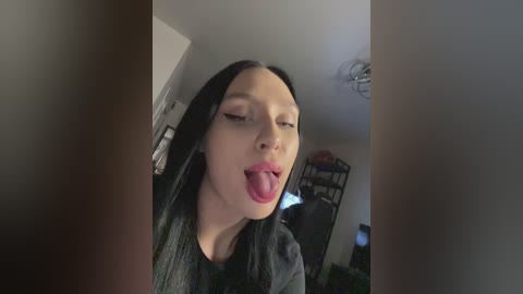 A video of a young woman with long black hair, light skin, and a playful expression, sticking out her tongue. She wears a dark top. The background shows a cluttered room with a shelf and a window.