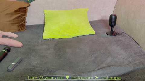 Media: Video of a gray cushion with a bright yellow pillow, a hand holding a green object, and a black microphone on a beige wall.