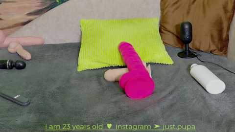 Media: Video of a woman lying on a grey bed with a neon green pillow, a black dildo, a pink dildo, and a white vibrator, in a cozy, dimly lit bedroom.