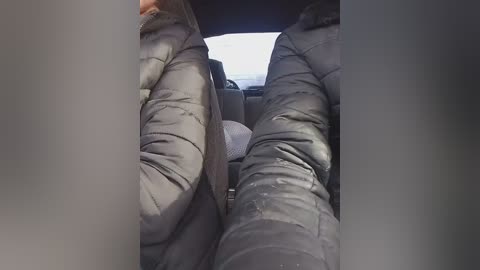 A video captures two people wearing dark, puffy jackets sitting in a car. The foreground features the jacket sleeves and hands. The background shows the car's interior with a blurry view of the road outside.