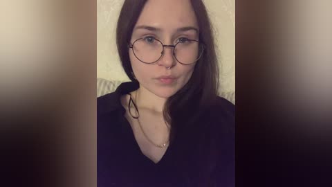 Video of a young woman with pale skin, straight brown hair, wearing round glasses and a black top, against a beige wall, taken indoors.