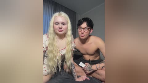 Media: Video of a blonde, slender woman in a floral top and a tattooed, shirtless man with glasses, sitting in a dimly lit room with gray curtains.