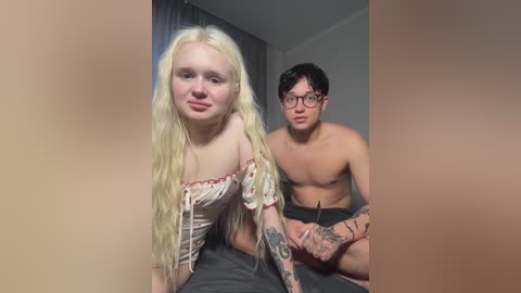 Media: Video of a blonde woman with fair skin, wearing a off-shoulder top, and a shirtless Asian man with glasses, both sitting indoors, blurry background.