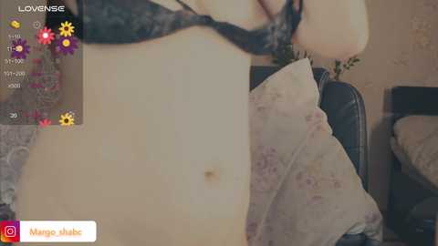 A video shows a light-skinned woman with a flat stomach, wearing a black lace bra, standing indoors. The background features floral pillows and a wooden table, with a watermark \"Manga Juke\" in the bottom left corner.